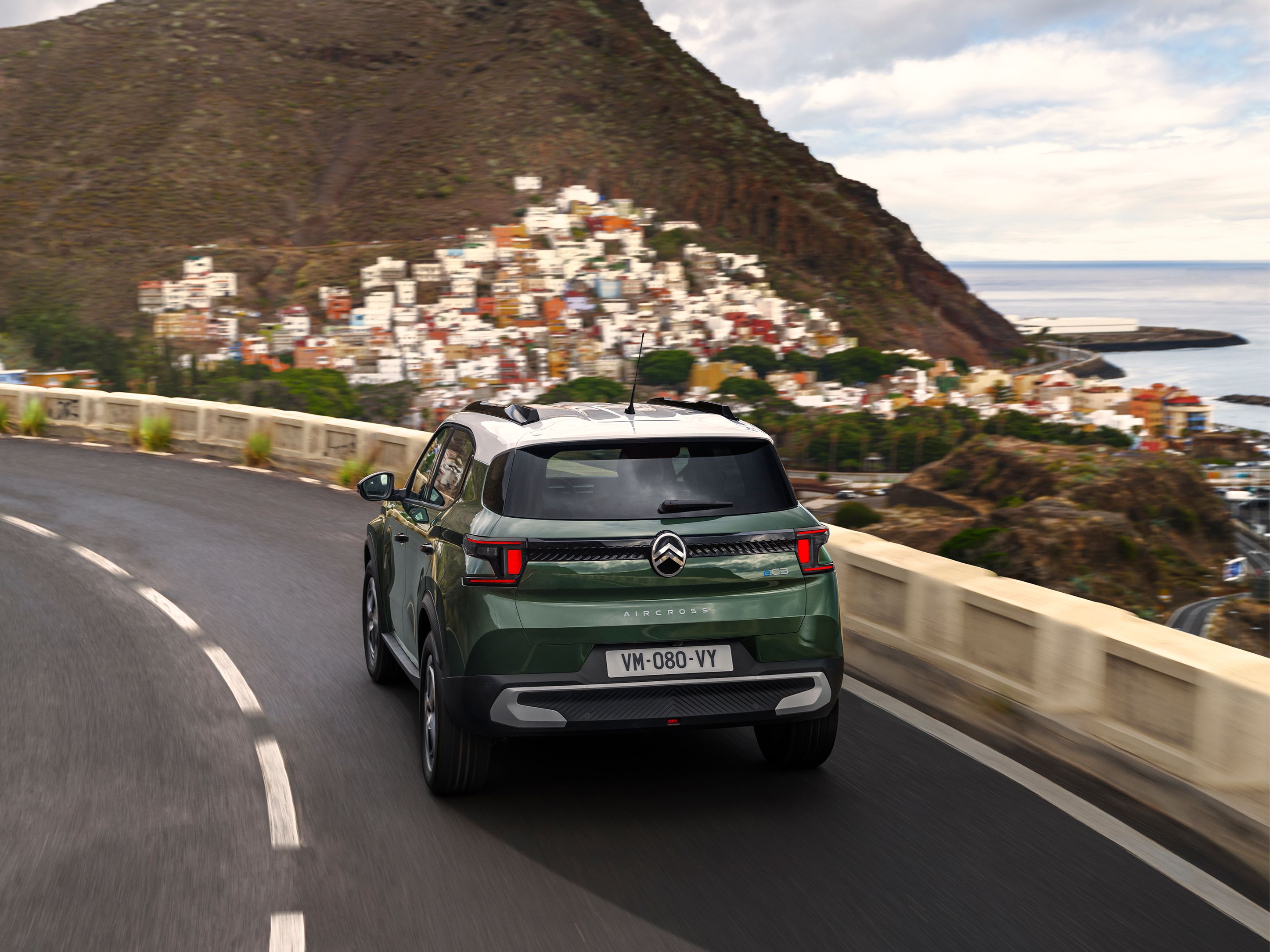 Dreiviertel-Heckansicht während der Fahrt des Citroën ë-C3 Aircross SUV auf einer Küstenstraße für sichere, sanfte und erschwingliche Alltagsmobilität.